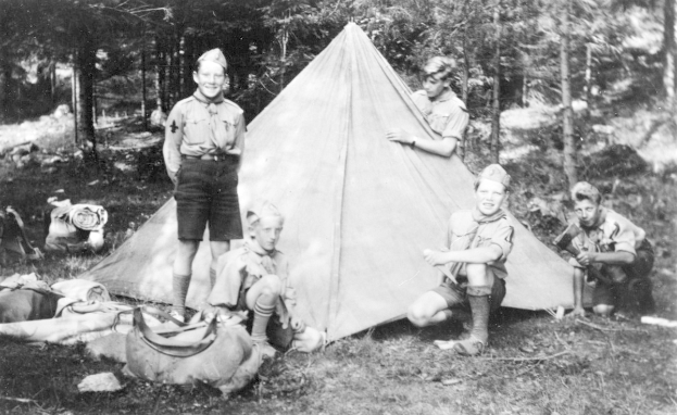 Schwarz-weiß-Foto von Pfadfindern in Uniformen, die sich um ein Zelt in einem bewaldeten Gebiet versammelt haben, einige sitzen und andere stehen, mit verstreuten Ausrüstungsgegenständen und Bäumen im Hintergrund.