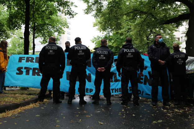Eine Gruppe von Polizeibeamten steht vor einer blauen Fahne mit Text, umgeben von Bäumen und Gebäuden in Berlin, Deutschland, mit einem geparkten Auto auf der rechten Seite und einer Frau in einer Jacke auf der linken Seite.