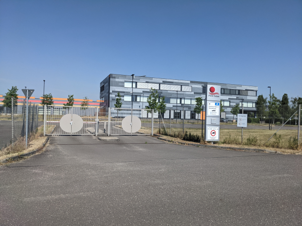Ein großes Gebäude mit vielen Fenstern, identifiziert als First Solar, ist von einem Metallzaun, Bäumen, Pflanzen, Gras, Schildern, Straßenlaternen und einem Weg umgeben.