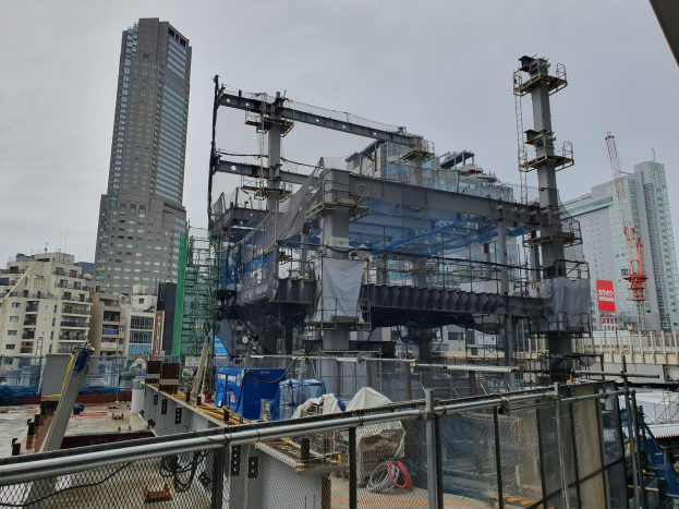 Großer Baustelle in einer Stadt mit einer teilweise gebauten Kraftwerkanlage, umgeben von einem Metallzaun, benachbarten Gebäuden, Versorgungsmasten, einem Banner, Fahrzeugen, öffentlichen Leitungen und einem bewölktem Himmel.