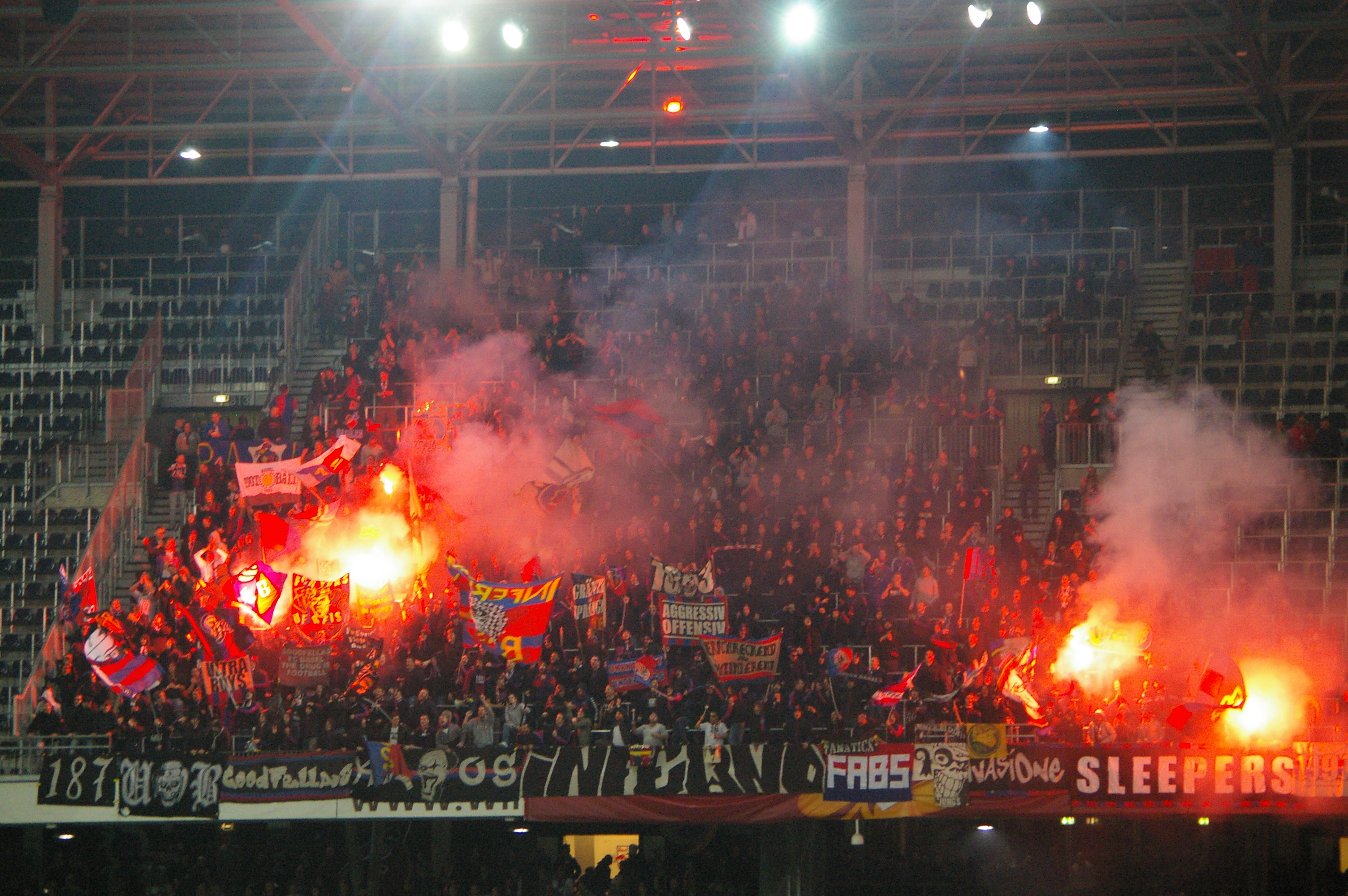 Eine große Menschenmenge in einem Stadion hält Fahnen und Banner, mit Leuchtraketen, die Rauch erzeugen, unter einem Dach mit Deckenlampen und Metallrahmen.