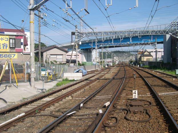 Elektrifizierte Bahnschiene mit Oberleitungen, eine Brücke, Gebäude, Bäume und einen klaren blauen Himmel.