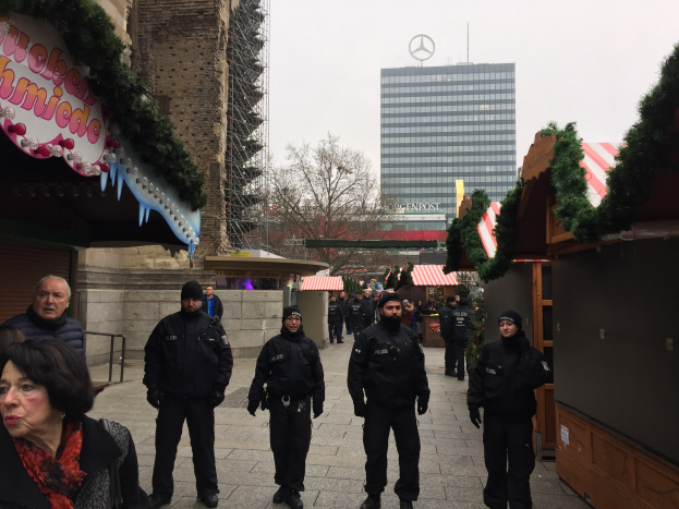 Polizisten in schwarzen Uniformen stehen vor einem geschmückten Weihnachtsmarkt in Berlin mit festlichen Dekorationen, Gebäuden, Bäumen und einem klaren blauen Himmel im Hintergrund.