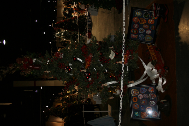 Ein geschm├╝ckter Weihnachtsbaum mit Geschenken, Rahmen und Spielzeugen auf einer Plattform, mit Menschen, weiteren Weihnachtsb├╝umen und festlichen Deko im Hintergrund.