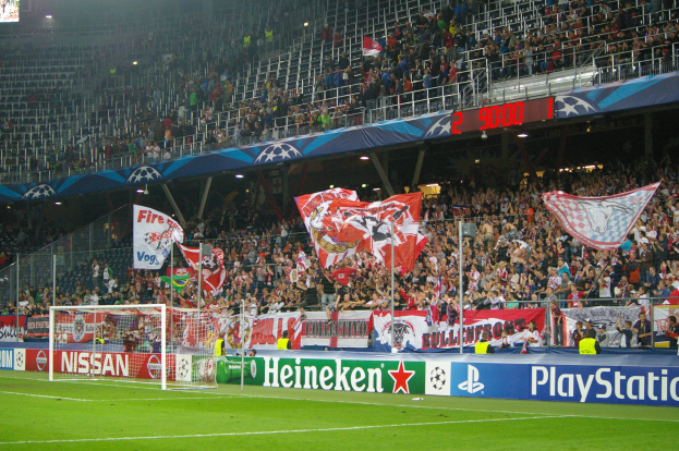 Ein Fussballspiel wird in einem hell erleuchteten Stadion mit einer grossen Menge, grünem Rasen, einem Torpfosten mit Bannern und einem Bildschirm in der oberen linken Ecke gespielt.