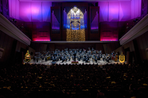 Großer Saal mit Publikum bei einem Konzert; auf der Bühne spielen Musiker Instrumente, während andere auf Stühlen sitzen; ein geschmückter Weihnachtsbaum und beleuchtete Wände sind im Hintergrund zu sehen.