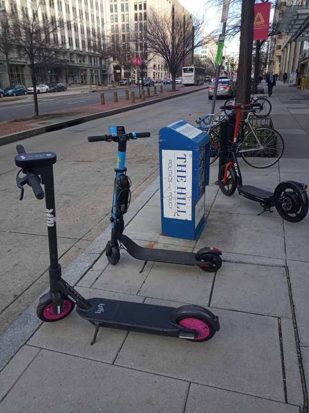 Zwei Elektro-Scooter auf einer städtischen Straße mit Fahrrädern, einem Müllcontainer, Fahrzeugen, Bäumen, Gebäuden und Menschen im Hintergrund geparkt.