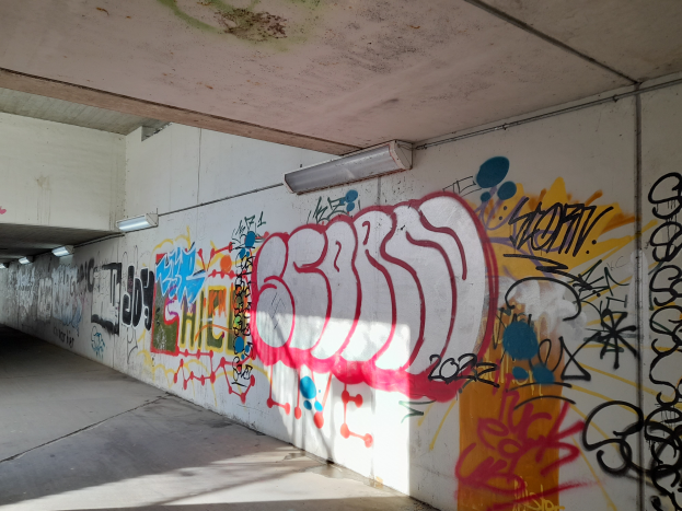Eine mit Graffiti bedeckte Wand in einer Tiefgarage, beleuchtet von Deckenlampen, mit einer Vielfalt an farbenfroher Straßenkunst.