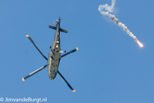 Ein Militärhubschrauber im Flug, der eine Rakete hält, die Rauch und Feuer ausstößt, mit Text in der unteren linken Ecke.