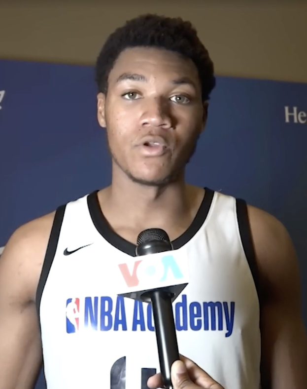 Ein Mann in einem weißen Tanktop hält ein Mikrofon und spricht mit der Medien an der NBA All-Star Game 2019, mit einem Banner und einer Wand im Hintergrund.