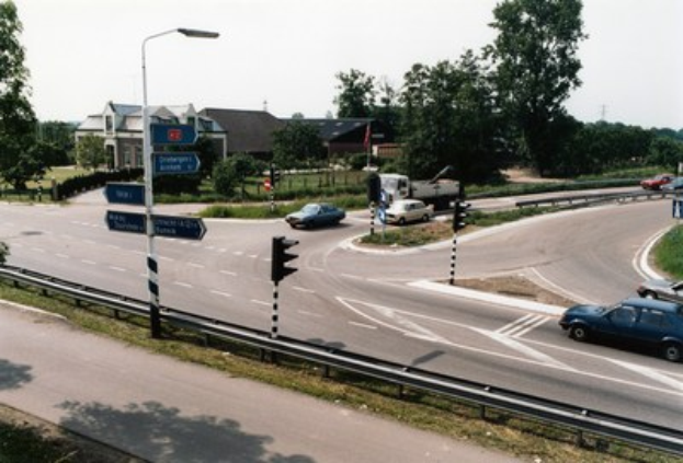 Kreisverkehr mit Autos, Ampeln, Verkehrszeichen, Laternen, Geländern, Grünflächen, Häusern und einem klaren blauen Himmel.
