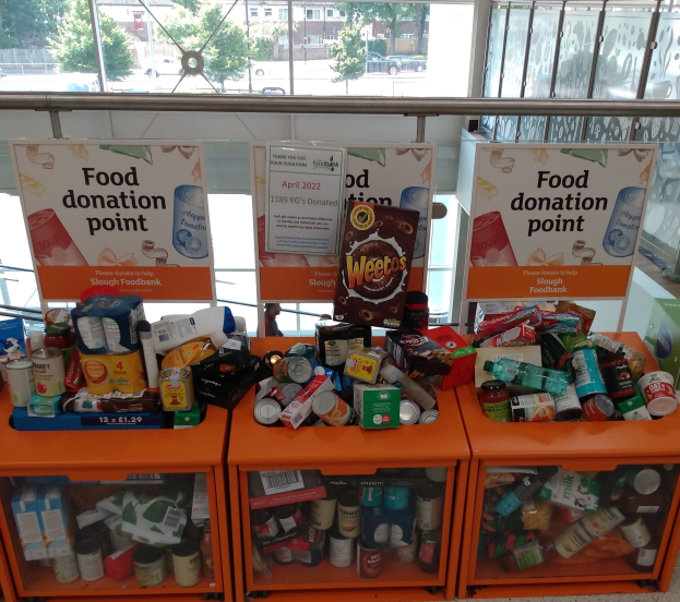 Orange Schränke gefüllt mit Lebensmittelspenden, darunter Flaschen, Dosen und Kartons, mit Informationsschildern und Blick durch Glasfenster auf Fahrzeuge, Bäume, Gebäude und Himmel.