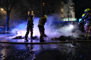 Feuerwehrleute in Helmen stehen nachts auf einer Straße mit Rauch, der aus einem Rohr auf der Straße aufsteigt, und halten Gegenstände, während Gebäude, Bäume und Fahrzeuge im Hintergrund zu sehen sind, mit einer Tafel rechts.