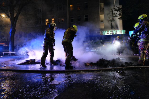 Feuerwehrleute in Helmen stehen nachts auf einer Straße mit Rauch, der aus einem Rohr auf der Straße aufsteigt, und halten Gegenstände, während Gebäude, Bäume und Fahrzeuge im Hintergrund zu sehen sind, mit einer Tafel rechts.