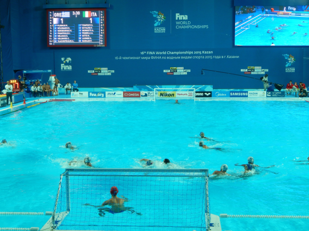 Eine Gruppe von Menschen, die Wasserball in einem großen Pool mit einem Netz in der Mitte spielen, Zuschauer auf der linken Seite und ein Bildschirm mit Text und Logos im Hintergrund bei den 2016 FINA Weltmeisterschaften in Kazan.