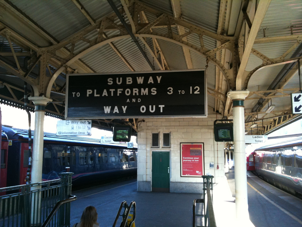 Zwei Züge stehen neben einem Bahnsteig mit zwei Bildschirmen und mehreren Schildern, die von der Decke hängen, eine Person ist in der Nähe einer Treppe und eine Tür ist an der Wand zu sehen.