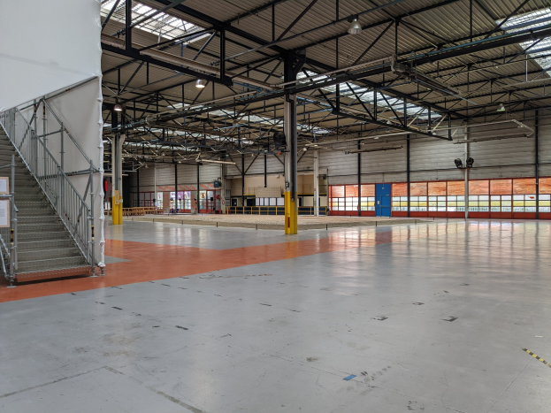 Großes Industriegebäude mit zahlreichen Fenstern, Metallstützen, einer Treppe mit Geländer, einem Schild, Deckenlampen und zusätzlichen Stützen.