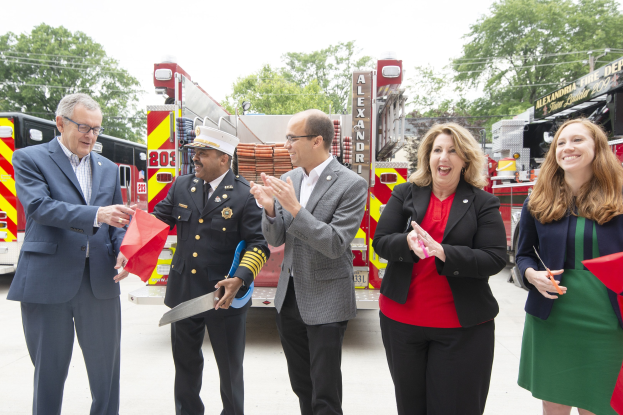 Group of people at a ribbon cutting ceremony for the Alexandria Fire Department, clapping and smiling in front of a fire truck, with two holding scissors and a red ribbon.
