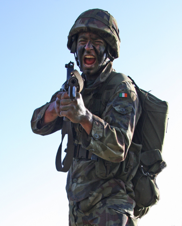 Eine Person in einer Armeeuniform, die ein Abzeichen, einen Helm und eine Tasche trägt und eine Waffe hält.