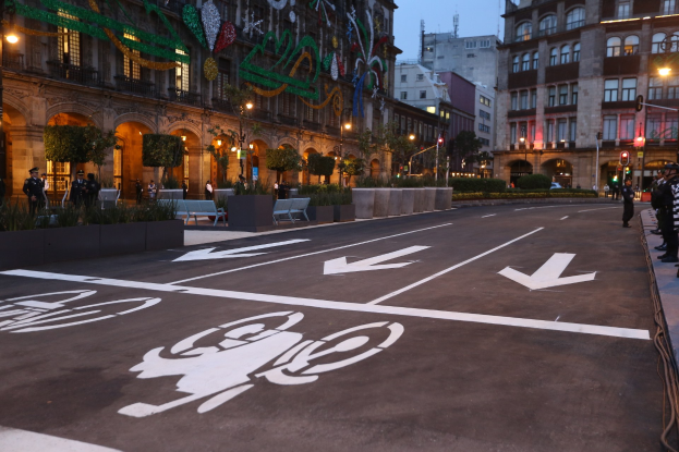Eine Stadtstraße mit einem ausgewiesenen Fahrradweg, gesäumt von Gebäuden, Straßenmöblierung, Fußgängern auf dem Gehweg, Grünflächen und einem klaren blauen Himmel.