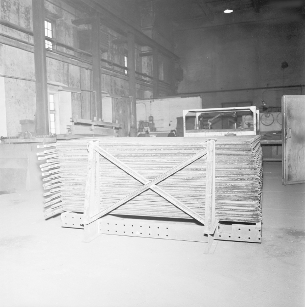 Ein Schwarz-Weiß-Foto einer Gießerei mit einem großen Metallgerüst, Metallobjekten auf dem Boden, einer Tür auf der rechten Seite, einer Wand mit Fenstern im Hintergrund und Lampen oben.