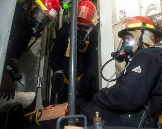 Drei Feuerwehrleute in Helmen und Gasmasken sitzen im Heck eines Trucks, tragen Uniformen und Handschuhe, mit einer Stange im Vordergrund und Rohren im Hintergrund, was auf einen begrenzten Raum hinweist.