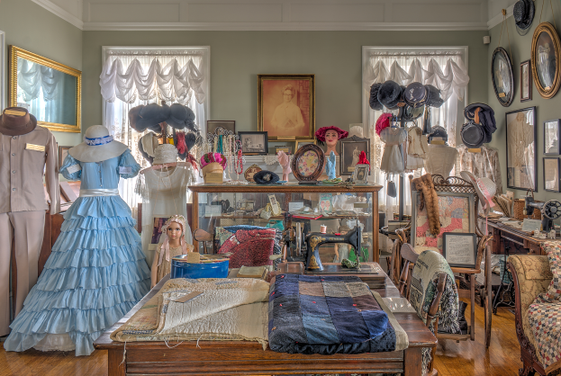 Ein Raum im Museum of Fine Arts in St. Louis, Missouri, voller Schaufensterpuppen in Kleidern und Hüten, ein Tisch mit Kleidung und einer Nähmaschine, Stühle mit Kissen, ein Schrank mit verschiedenen Gegenständen, Fotorahmen an der Wand, Fenster mit Vorhängen und einer Decke.