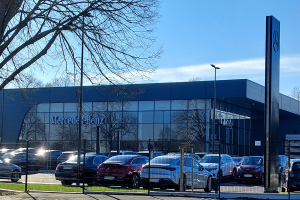 Ein Mercedes-Autohaus mit parkenden Autos davor, umgeben von einem Metallzaun, Gras, einem Schild, Straßenlaternen, Bäumen und einem bewölkten Himmel.