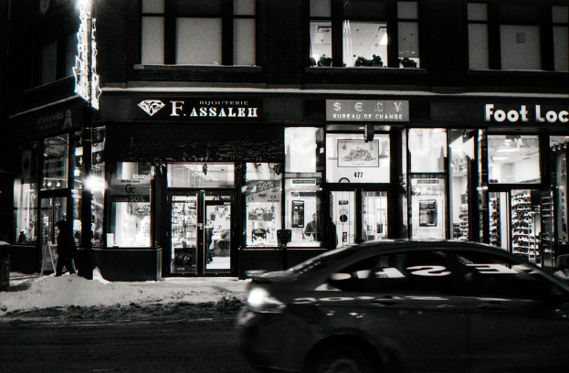 Ein Schwarz-Weiß-Bild eines Autos auf einer Straße mit einem Gebäude mit Glasfenstern oben und Lagerbereichen darunter, mit Werbetafeln in der Nähe der Geschäfte und einer Lampe auf der linken Seite.