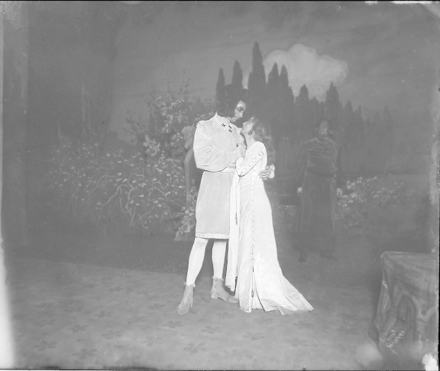Ein Schwarz-Weiß-Foto eines Mannes und einer Frau, die sich auf einer Bühne umarmen und küssen, mit Menschen, Pflanzen und Bäumen im Hintergrund.