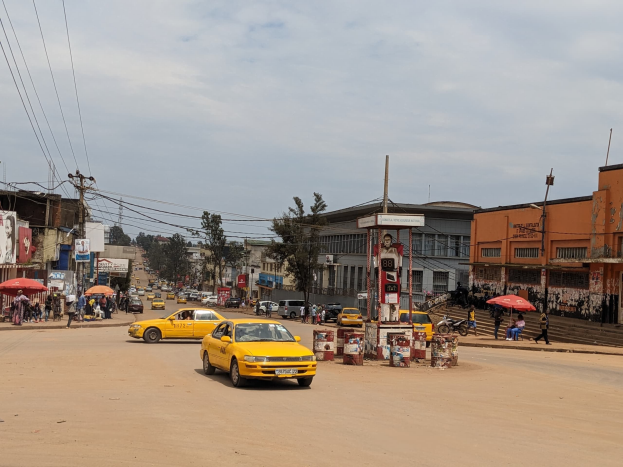 Ein gelbes Taxi fährt auf einer belebten Stadtstraße mit Gebäuden an beiden Seiten, während Fußgänger mit Regenschirmen auf dem Gehweg gehen, mit Strommasten und Kabeln im Hintergrund.