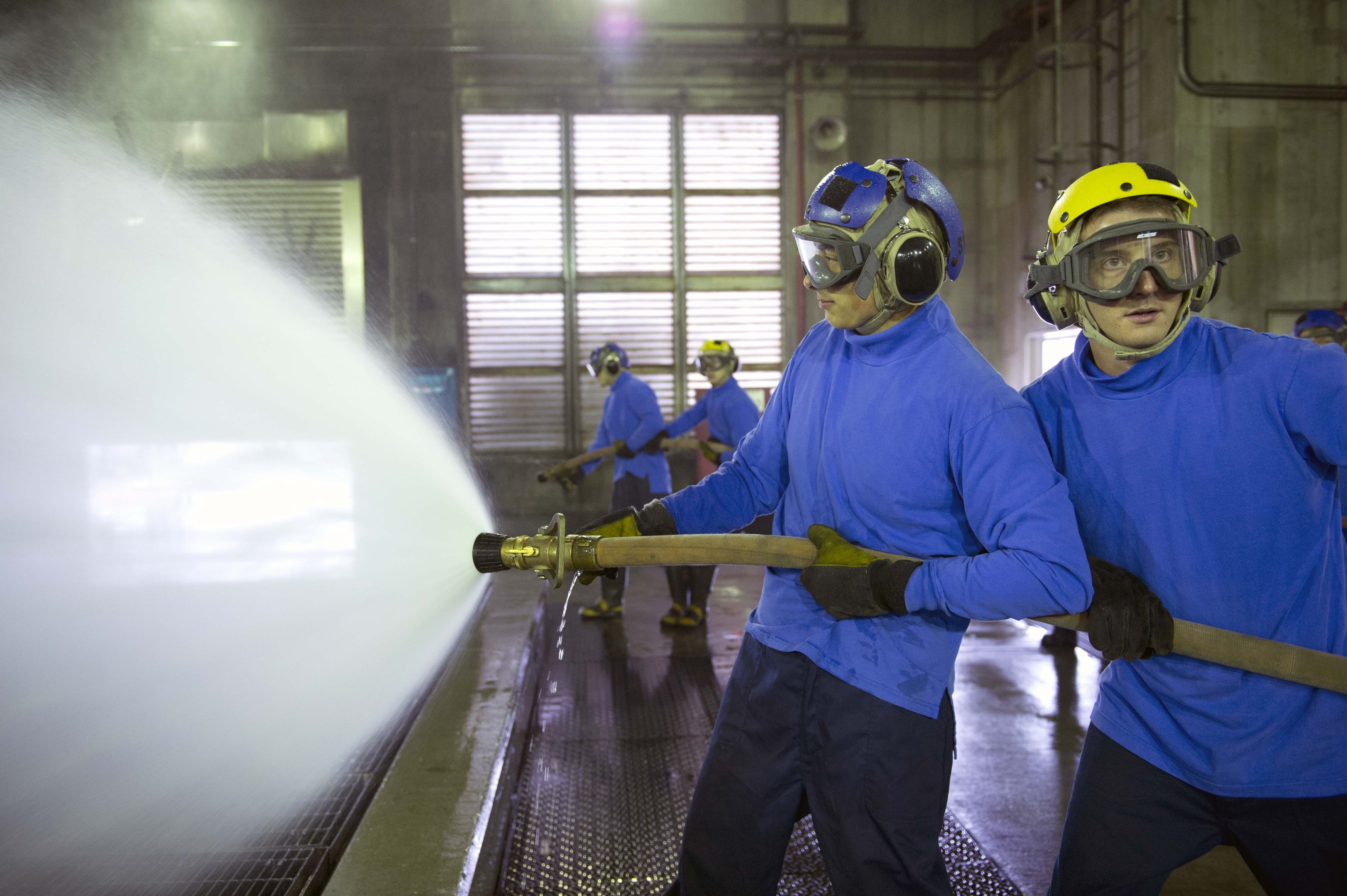 Gruppe von Männern in blauen Hemden und gelben Helmen, die an Maschinen arbeiten, einer sprüht Wasser auf den Boden in einer Fabrikumgebung mit sichtbaren Rohren, Fenstern und Lichtern.
