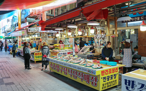 Ein belebter Straßenmarkt in Seoul, Südkorea mit Menschen, Ständen mit verschiedenen Gegenständen und Gebäuden unter einem klaren blauen Himmel.