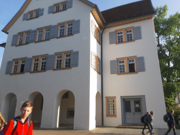 Eine Gruppe von Menschen, einige mit Taschen, geht vor einem großen weißen Gebäude mit Fenstern und Türen, umgeben von Bäumen unter einem klaren blauen Himmel, vorbei, was darauf hindeutet, dass sie auf dem Weg zu einer Schule in Deutschland sind.