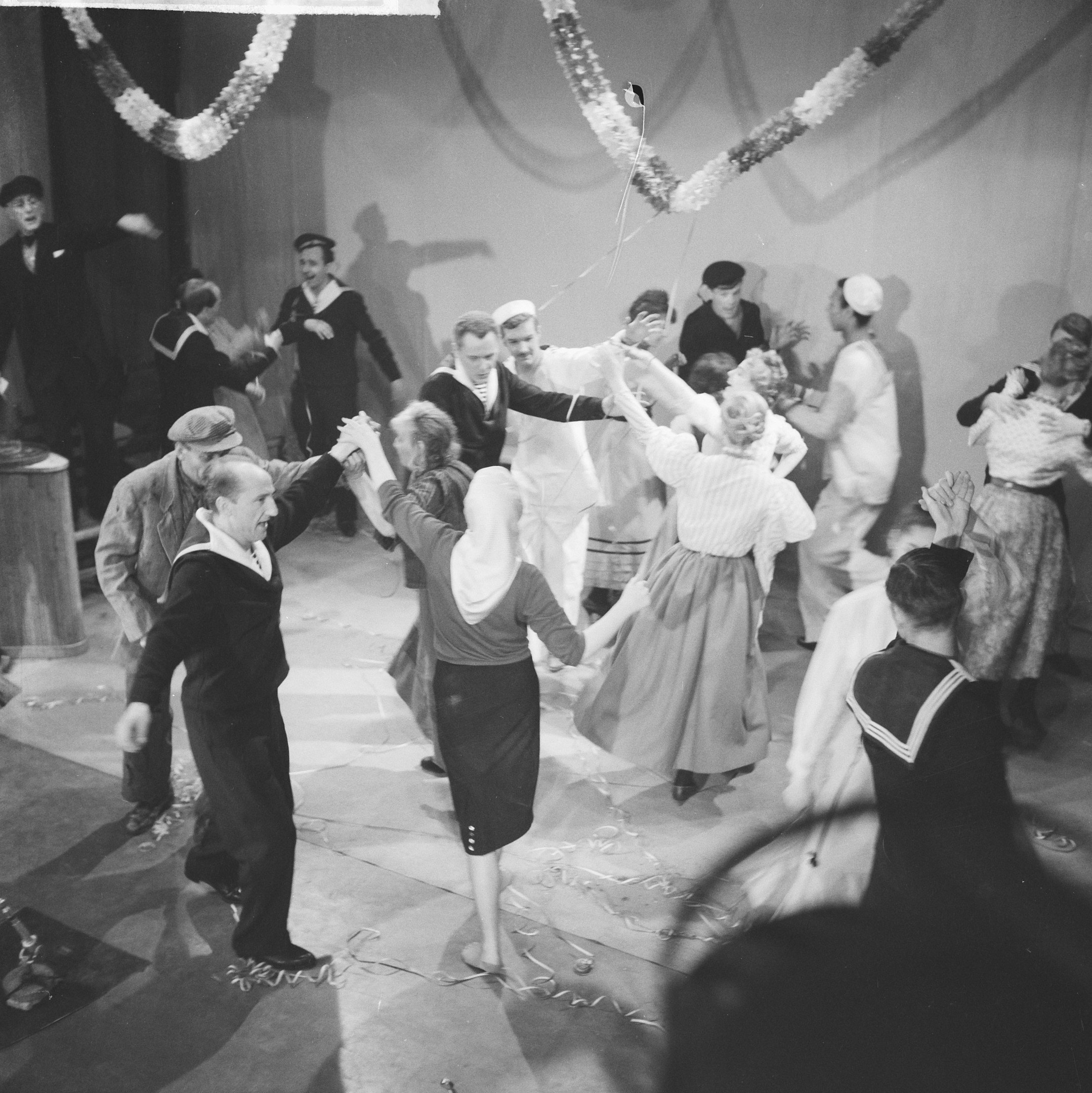 Schwarz-weiß-Bild von Menschen, die auf einer Tanzfläche tanzen, mit einer dekorierten Wand im Hintergrund und einem Tisch mit Gegenständen links daneben.