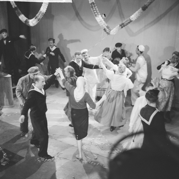 Schwarz-weiß-Bild von Menschen, die auf einer Tanzfläche tanzen, mit einer dekorierten Wand im Hintergrund und einem Tisch mit Gegenständen links daneben.
