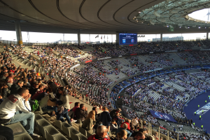 Eine große Menschenmenge sitzt in einem Stadion und schaut ein Fußballspiel, mit einer Bühne rechts daneben, Fahnen, Stangen und einem Bildschirm im Hintergrund, unter einem sichtbaren Himmel, im Allianz Arena in München, Deutschland.