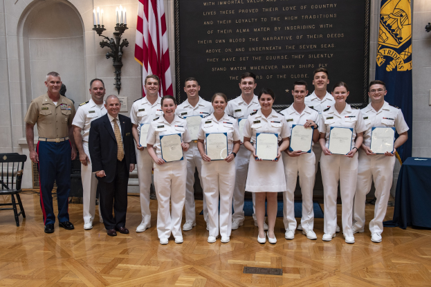Gruppe uniformierter Personen, die strahlend mit Urkunden in der Hand posen, neben einem blauen Tisch und Stühlen, mit Fahnen, Lichtern und einer Tafel mit der Aufschrift "U.S. Naval Academy Class of 2018" im Hintergrund.