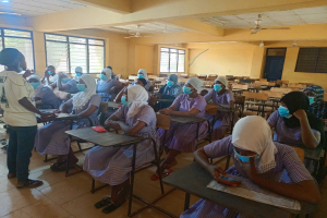Ein Klassenzimmer in Nigeria mit Schülern, die Gesichtsmasken tragen und auf Bänken und Stehplätzen sitzen, mit Büchern und Gegenständen auf den Tischen, Fenstern, einer Tür und Deckenleuchten und Ventilatoren im Hintergrund.