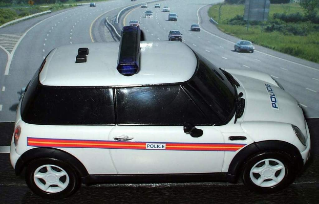 Weißer Polizeiwagen mit "Polizei" Aufschrift auf einer Vorstadtstraße, umgeben von Verkehr, Gras und Bäumen auf beiden Seiten.