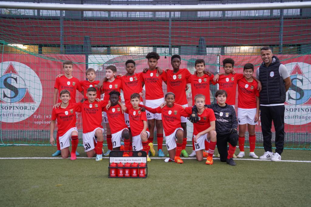 Eine Gruppe junger Männer in roten T-Shirts und schwarzen Shorts steht auf einem Fußballfeld, mit einer Kiste roter Gegenstände vor ihnen und Bannern mit 'FC Red Black Eglaute - Champions League' im Hintergrund.