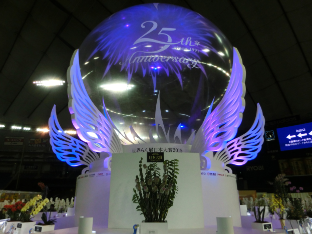 Ein großer Kristallball mit Flügeln oben drauf, umgeben von Blumensträußen, Namensschildern und anderen Gegenständen auf dem Boden, mit Tafeln, Lichtern und einem Bildschirm im Hintergrund bei einer 25-jährigen Jubiläumsfeier.