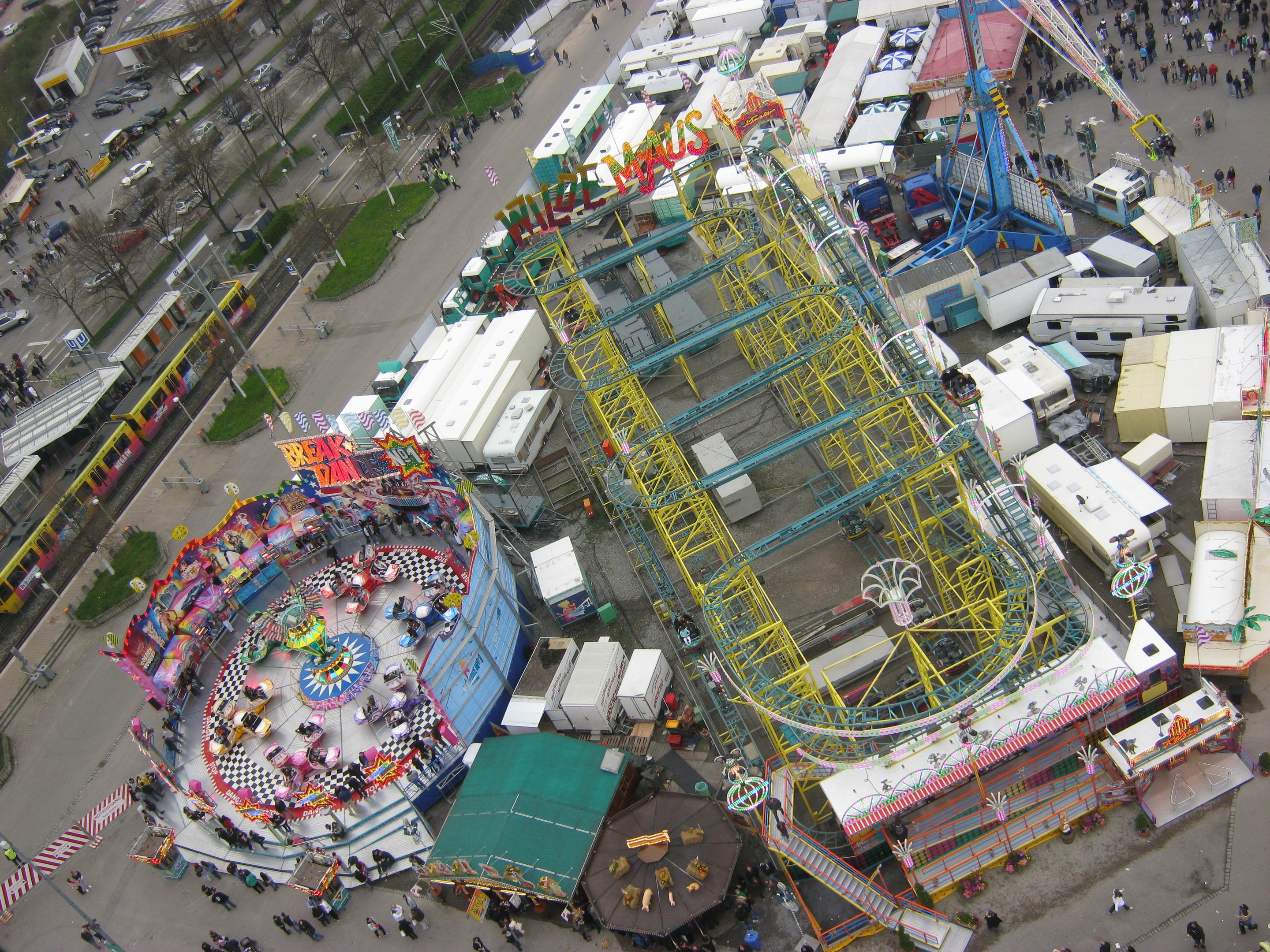 Luftaufnahme eines Vergnügungsparks mit einem zentralen Riesenrad, umgeben von verschiedenen Attraktionen, Gebäuden, Fahrzeugen, Menschen, Wegen und Grünflächen.