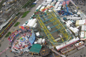 Luftaufnahme eines Vergnügungsparks mit einem zentralen Riesenrad, umgeben von verschiedenen Attraktionen, Gebäuden, Fahrzeugen, Menschen, Wegen und Grünflächen.