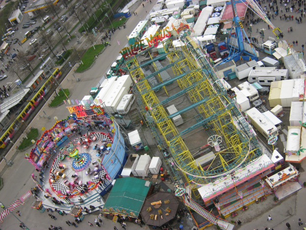 Luftaufnahme eines Vergnügungsparks mit einem zentralen Riesenrad, umgeben von verschiedenen Attraktionen, Gebäuden, Fahrzeugen, Menschen, Wegen und Grünflächen.