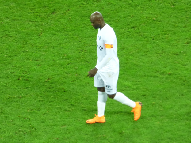 Ein Füßballspieler in einem weißen T-Shirt, weißen Shorts und orangefarbenen Schuhen überquert ein saftig grünes Feld.