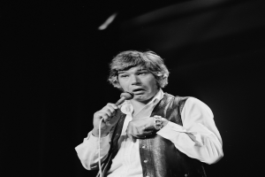 Ein Mann in weißem Hemd und schwarzer Weste singt leidenschaftlich in ein Mikrofon, seine Augen sind geschlossen und seine Haare sind im klassischen Rocker-Stil frisiert.