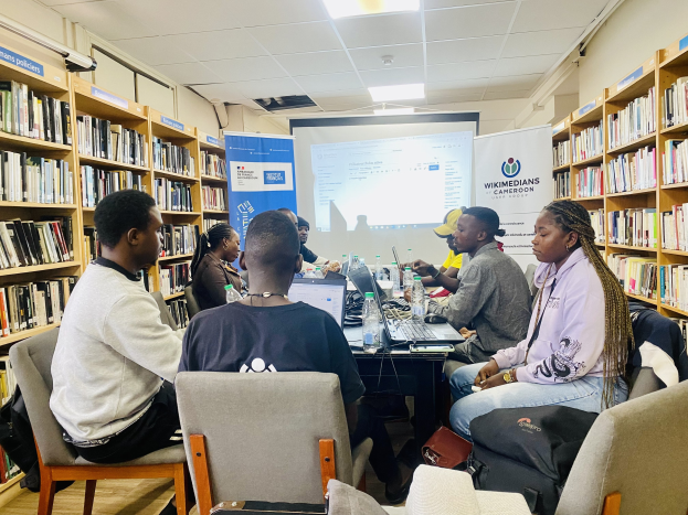 Gruppe von Menschen, die um einen Tisch in einer Bibliothek sitzen und diskutieren, mit Laptops und Gegenständen auf dem Tisch, umgeben von Bücherregalen und Bannern im Hintergrund.