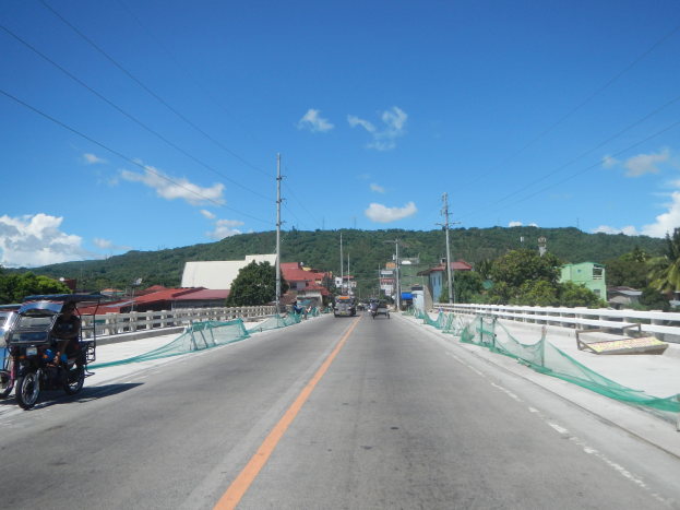 Autobahn mit fahrenden Fahrzeugen, ein geparktes Motorrad auf dem Seitenstreifen, Geländer, Strommasten mit Drähten, Bäume, Gebäude, Hügel und ein bewölkter Himmel im Hintergrund.