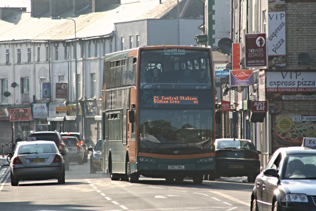 Eine Straße mit Autos und einem Bus im Vordergrund, mit Gebäuden mit Wänden, Fenstern, Tellern und Dächern im Hintergrund, sowie Plakaten und Bannern an den Wänden und einem Laternenmast.